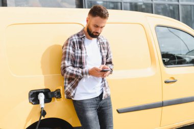 Elektrikli minibüsü şarj olurken akıllı telefon kullanan kargo şoförü sürdürülebilir ulaşım ve modern lojistiği temsil ediyor.