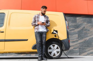 Sarı elektrikli minibüsü modern binanın önünde şarj olurken teslimatçı akıllı telefon kullanıyor.