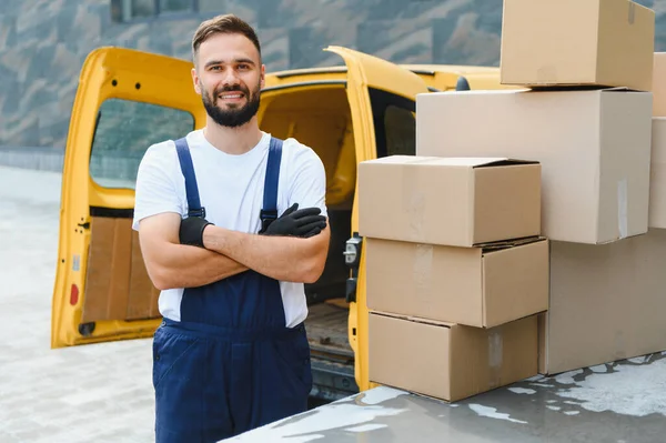 Eldiven ve üniforma giyen kurye, kolları çaprazlanmış bir şekilde karton kutular ve sarı bir minibüsün yanında gülümsüyor.