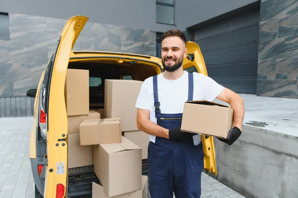Kurye gülümsüyor ve karton kutularla dolu sarı minibüsünün yanında bir paket tutuyor.