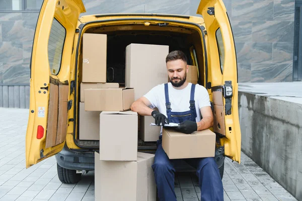Eldiven takan kurye, karton kutularla dolu sarı minibüste oturuyor. Dosyada nakliye listesini kontrol ediyor.