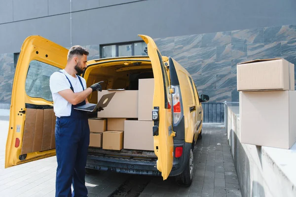 Eldiven takan kurye, bir dizüstü bilgisayarda çalışırken şehir caddesinde sarı bir minibüsün içindeki karton kutuları kontrol ediyor.