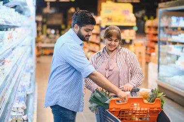 Mutlu Hintli çift canlı bir süpermarkette alışveriş arabasını itiyor, taze yiyecek seçiyor ve birlikte vakit geçiriyorlar.