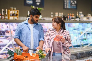 Genç Hintli çift alışveriş arabasını itiyor ve kalabalık bir markette birlikte vakit geçirirken paket et seçiyorlar.