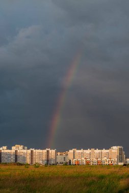 Fırtınalı bir gökkuşağı ve gökkuşağına karşı, güneşin altın ışınlarında yüksek binalar kompleksi. Şehrin üzerinde gün batımı günü.