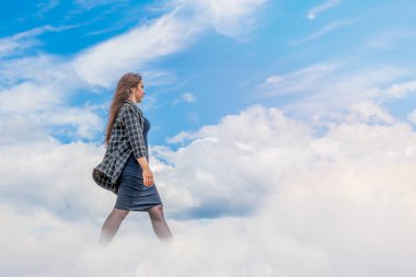 Genç bir kız yavaşça bulutların arasında yürüyor koyu bir elbise ve mavi bir arka planda kareli bir ceketle.