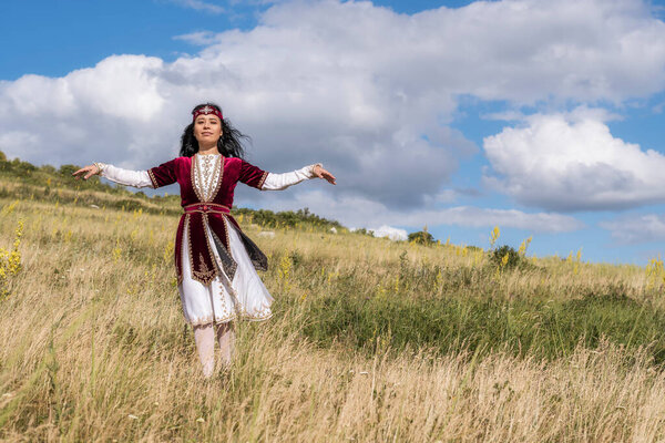 A young bright girl in a beautiful red suit is dancing a national dance on the field against a blue summer sky