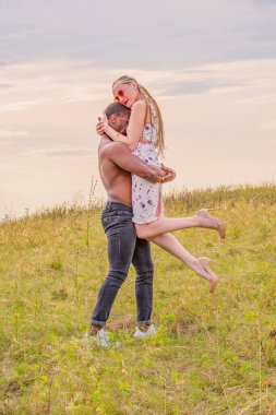 Koyu tenli genç bir adam kollarında beyaz bir kızı kaldırdı ve yeşil bir tarlanın ve gün batımının arka planında birbirlerine tutundular.