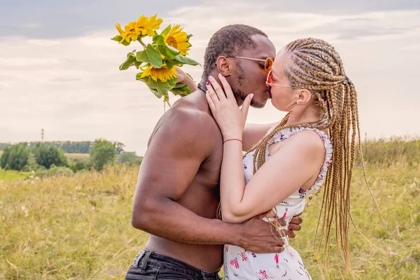 A dark-skinned young guy froze a girl in a gentle kiss against the background of a green field and a summer evening sky. Girl holding a bouquet of yellow sunflowers in her hands