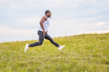 Genç bir adam yeşil bir çayırda koşuyor ve engellerle koşarken yükseğe zıplıyor.