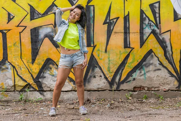 A young, happy girl in a denim suit and a bright T-shirt stands against the background of a wall with a blue graffiti pattern and smiles