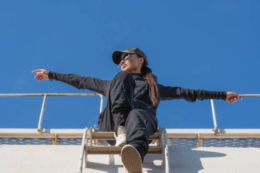 Siyah takım elbiseli ve aynalı gözlüklü genç bir kız mavi gökyüzüne karşı kollarını yana açmış bir tank vagonunun merdivenlerinde oturuyor.