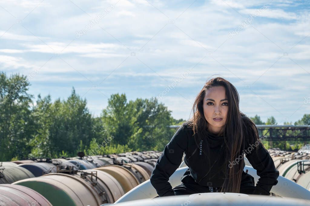 A young girl in a black suit gazes ahead. In the background there are railway tanks and a blue sky.