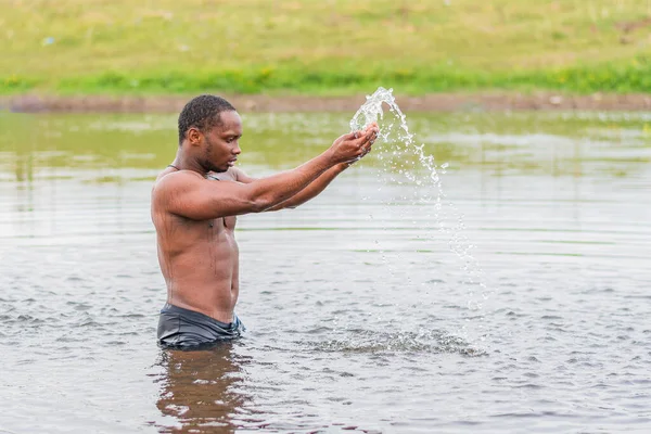 A young guy of athletic build stands waist-deep in the water of the lake, raises splashes with his hands and looks at them