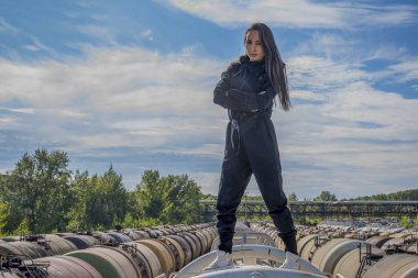 Siyah takım elbiseli genç bir kız, kolları göğsünde, mavi gökyüzünün arka planında katlanmış metal bir yüzeyde duruyor.