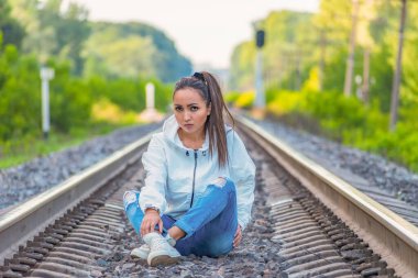 Güzel, genç ama yorgun ve üzgün bir kız beyaz ceketli ve kot pantolonlu rayların arasında oturuyor ve kameraya bakıyor. Tek bir turistin yaz maceraları.