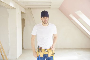 Genç tamirci inşaat alanında duran beyzbol kap ve aracı kemer takmaya portresi.