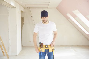 Genç tamirci inşaat alanında duran beyzbol kap ve aracı kemer takmaya portresi.