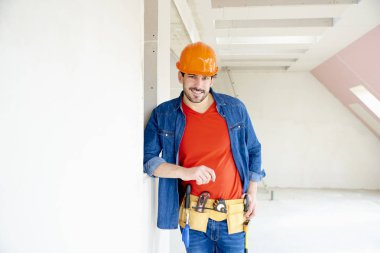 Emanet kask ve alet kemeri takıyor ve inşaatta çalışırken çekiç matkap sürücüsünü kullanan genç tamirci portresi. 