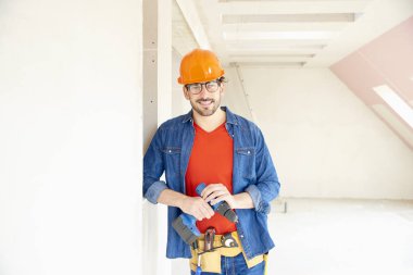 Emanet kask ve alet kemeri takıyor ve inşaatta çalışırken çekiç matkap sürücüsünü kullanan genç tamirci portresi. 