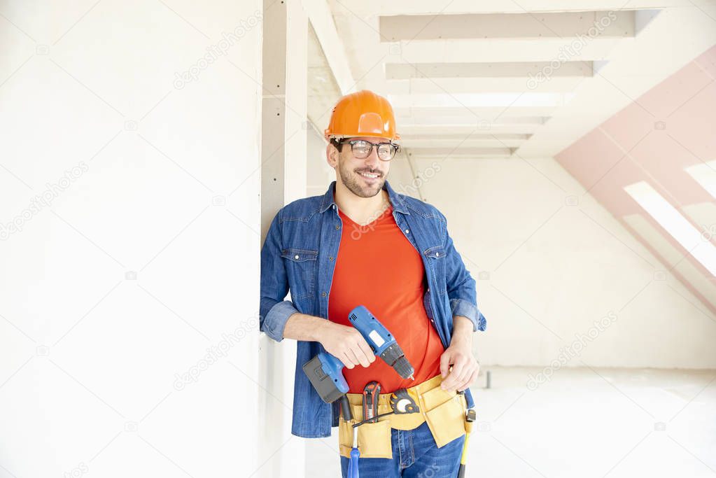 Portrait of young repairman wearing safety helmet and tool belt and using hammer drill driver while working on construction site. 