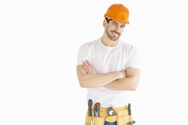 Portrait of young handyman wearing hard hat and tool belt while standing with arms crossed at isolated white background. 