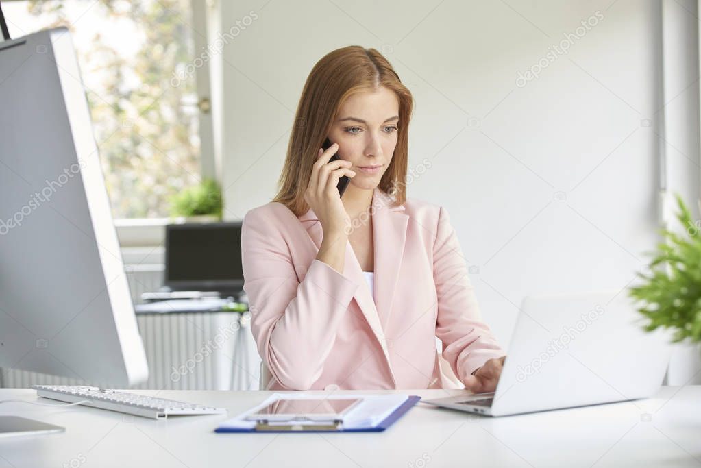 Attractive young businesswoman making call and using laptop while working at the modern office. 
