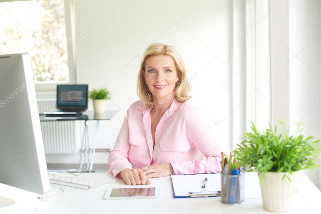 Smiling beautiful sales woman sitting at office desk while using digital tablet and computer. 