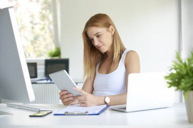 Ofiste çalışan gülümseyen genç bir kadın. Güzel iş kadını ofis masasında oturuyor ve dijital tablet kullanıyor..