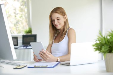Ofiste çalışan gülümseyen genç bir kadın. Güzel iş kadını ofis masasında oturuyor ve dijital tablet kullanıyor..