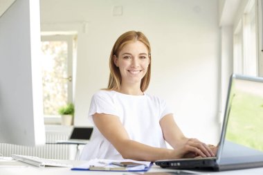 Ofis masasında otururken ve internette çalışırken gülümseyen genç iş kadınının fotoğrafı.. 