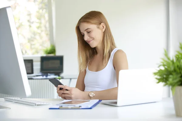 彼女の携帯電話とテキスト メッセージング Iat のオフィスの机に座っていると作業しながら使用して美しい若い実業家 — ストック写真