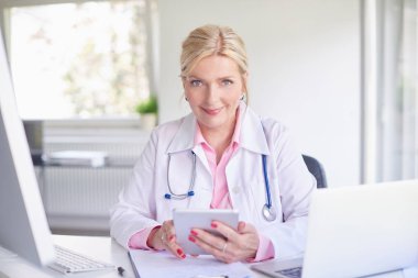 Üst düzey kadın doktor doktor odasında dizüstü bilgisayar ve bilgisayar önünde otururken Touchpad kullanarak