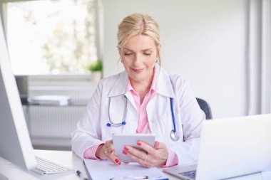 Üst düzey kadın doktor doktor odasında dizüstü bilgisayar ve bilgisayar önünde otururken Touchpad kullanarak