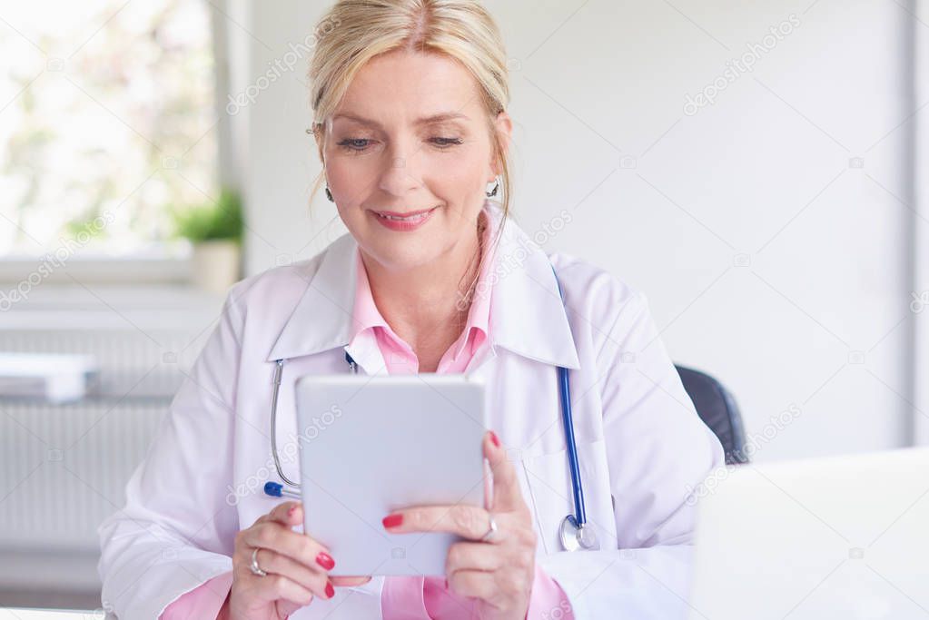 Senior female doctor using touchpad while sitting at doctors room in front of laptop and computer