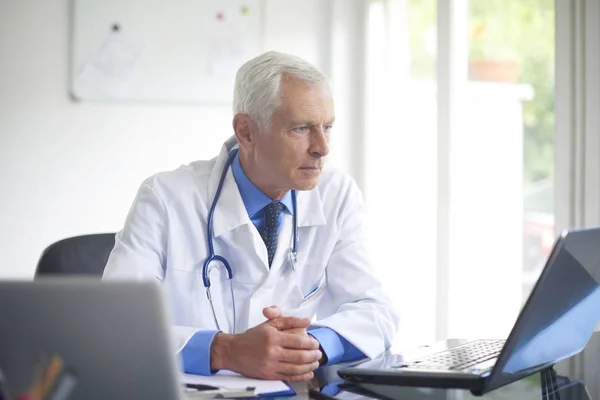 Portrait of senior male doctor working on laptop while sitting in consulting room.