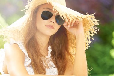 Güzel genç kadının açık ve kamera bakarak otururken hasır şapka ve güneş gözlüğü takıyor headshot.