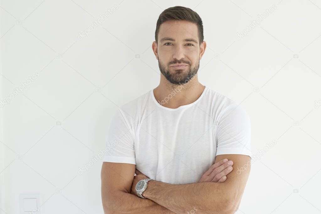 Studio portrait of arms crossed young man wearing white t shirt while looking at camera and smiling. Isolated on white background. 