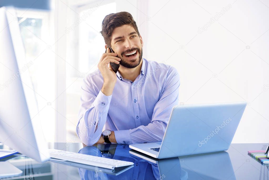 Happy businessman wearing shirt while sitting at office desk behind his laptop and making a call. 