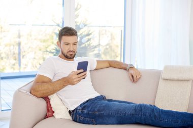 Yakışıklı genç adam cep telefonunu kontrol ediyor ve evdeki kanepede dinlenirken mesajlar okuyor.. 
