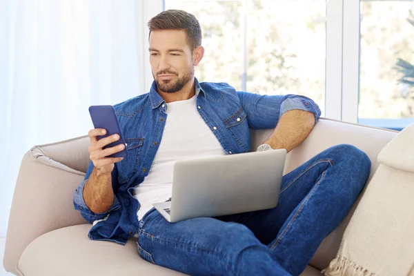 Portrait shot of a happy young man wearing casual clothes while using laptop and text messaging on sofa at home.