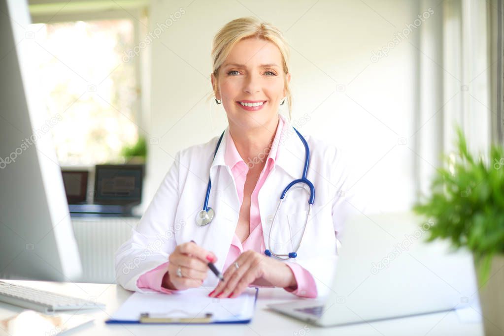 Beautiful smiling female doctor sitting in front of her computer at the doctor's office and writing something. 