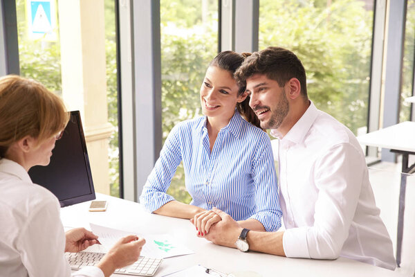 Financial advisor businesswoman sitting at office desk in front of computer and consulting with young couple about finance tips.