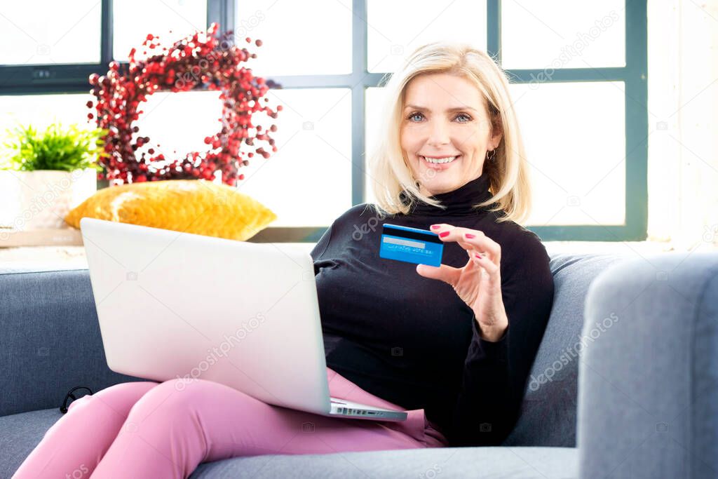 Portrait shot of attractive senior woman holding bank card in her hand and using laptop while shopping online. 
