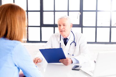 Orta yaşlı erkek doktorun, doktorların odasında masasında otururken hastasıyla konuşması.