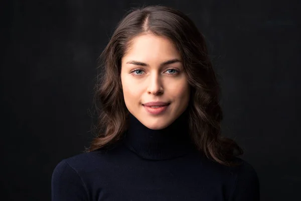 Headshot of attractive young woman with beautiful smile wearing turtleneck sweater while posing at dark background.