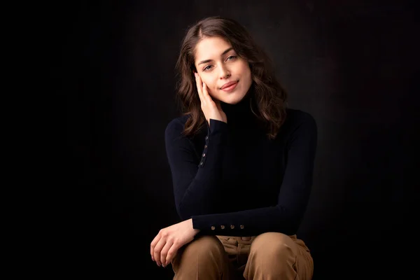 Portrait of beautiful woman wearing turtleneck sweater and smiling while sitting at isolated dark background.