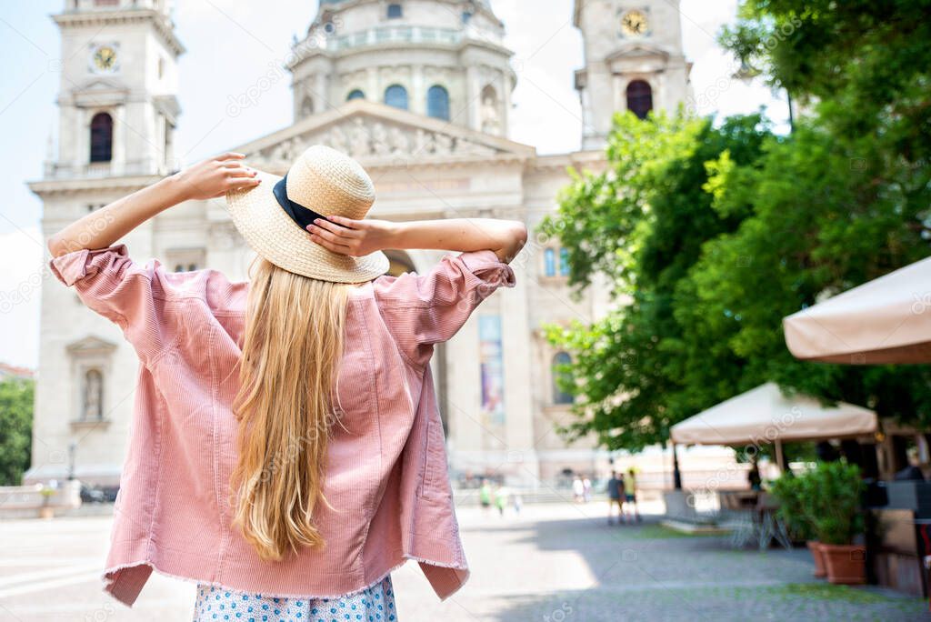Rear view shot of long haired woman wearing straw hat while standing with arms outstretched in front of St. Stephen Basilica in Budapest.