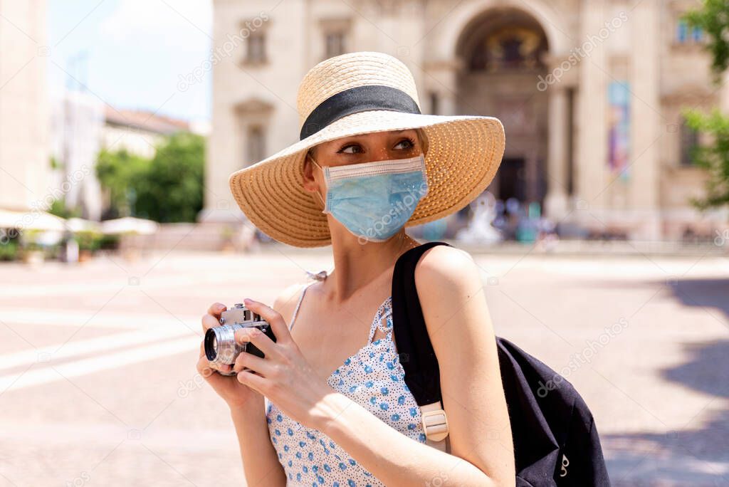 Portrait shot of beautiful blond woman wearing face mask for prevention while using camera to taking photos in the city. 