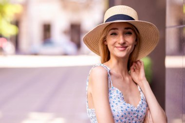 Sokakta dikilirken hasır şapka ve yaz elbisesi giyen çekici genç bir kadının yakın plan portresi.. 
